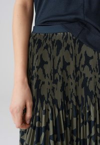 Gonna plissettata con stampa animale verde e blu navy, con una cintura solida blu navy. Mano che afferra il tessuto lungo la cucitura.