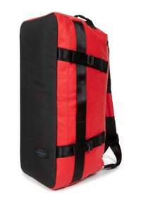 Sac à dos en tissu rouge et noir de forme rectangulaire, avec des bretelles ajustables et des accents en matériel noir. Présente des boucles latérales robustes et un patch logo.