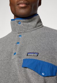 Grijze fleece pullover met half ritssluiting, voorzien van blauwe knopen en kraagaccenten; met een voorzak en het Patagonia-logo op de borst.