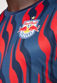 Piros és kék mintázatú sportme, kerek nyakkivágással. Szerepel rajta a Red Bull Salzburg logója és egy texturelt szövetdesign.