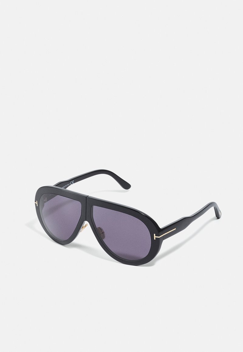 Occhiali da sole neri con montatura oversize e angolare, lenti viola scure e dettagli in tono oro sulle aste. Finitura opaca con una texture liscia.