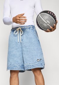 Lyseblå denimshorts med elastisk talje og hvid snor, prydet med et sort Nike-logo på venstre ben, der holdes med en basketball.