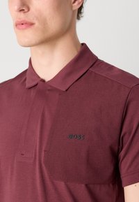 Polo tričko s krátkym rukávom v burgundskej farbe s textúrovanou nášivkou na hrudi a čiernym logom "BOSS". Vyrobené z mäkkého, priedušného materiálu.