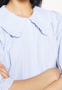 Chemise rayée bleu clair et blanche avec un col volanté, des boutons sur le devant et des manches courtes. Le tissu semble doux et texturé.