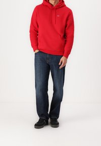 Sudadera roja con capucha y bolsillo tipo canguro, combinada con vaqueros azul oscuro de pierna recta y zapatillas negras. Diseño sencillo, tejido cómodo.