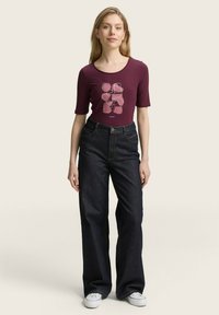 Bordeauxfarbener kurzärmeliger Body mit abstraktem Grafikdesign, kombiniert mit dunkelblauen weiten Jeans und weißen Sneakers vor einem neutralen Hintergrund.