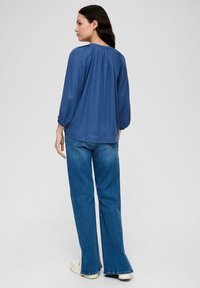 Blouse bleu ample à manches trois-quarts, avec un dos froncé, associée à un jean en denim évasé et des baskets blanches.