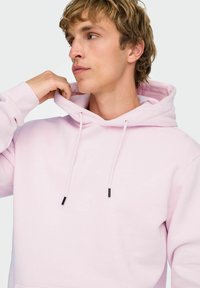 Felpa con cappuccio rosa chiaro con tasca frontale, dettagli con cordoncini neri e polsini a costine. Texture liscia, vestibilità comoda.