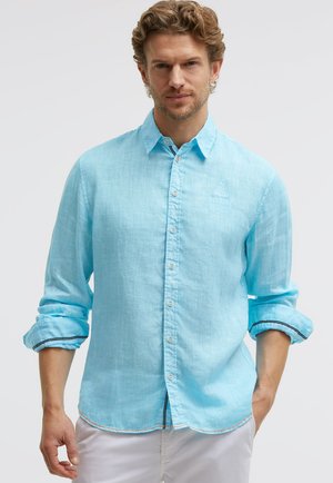 Mann, der ein hellblaues langärmliges Hemd mit hochgekrempelten Ärmeln und eine weiße Hose trägt, steht mit einer Hand in der Tasche.