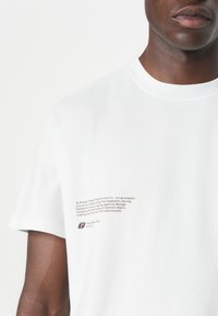 Weißes Baumwoll-T-Shirt mit kurzen Ärmeln und rundem Ausschnitt. Auffälliger brauner Text auf der linken Seite mit einem Logo und Branding-Details.