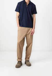 Mörkblå kortärmad polo med dragkedja i kragen, i kombination med beige löst sittande byxor och bruna robusta snörskor.