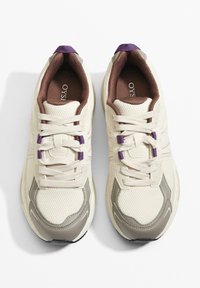 Krémové tenisky s horní částí z síťoviny, polstrovaným hnědým límcem, šněrováním vpředu, šedými detaily po stranách a fialovými poutky na tkaničkách. Plochá černá podrážka.