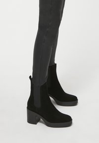 Botines de ante negros con tacón cuadrado y paneles laterales elásticos. Textura suave con punta redondeada y una lengüeta en la parte trasera.