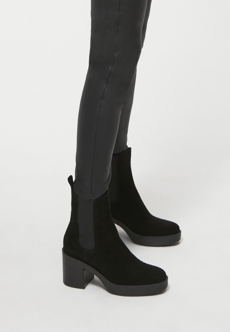 Botines de ante negros con tacón cuadrado y paneles laterales elásticos. Textura suave con punta redondeada y una lengüeta en la parte trasera.