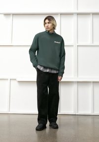 Grøn crewneck sweatshirt med et logo, båret over en sort ternet skjorte. Sammen med sorte corduroy bukser med brede ben og sorte sneakers.