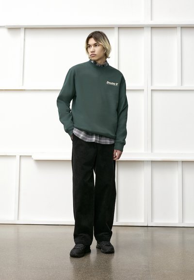 Sweatshirt vert à col rond avec un logo, porté par-dessus une chemise à carreaux noire. Assorti à un pantalon large en velours côtelé noir et des baskets noires.