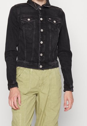 Person, die eine taillierte schwarze Jeansjacke mit silbernen Knöpfen und hellgrünen Cargo-Hosen trägt, steht vor einem schlichten Hintergrund.