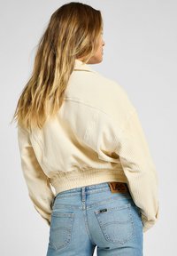 Chaqueta de pana beige recortada con textura acanalada y cintura elástica, llevada con jeans de mezclilla azul claro que tienen bolsillos traseros y etiqueta de marca.