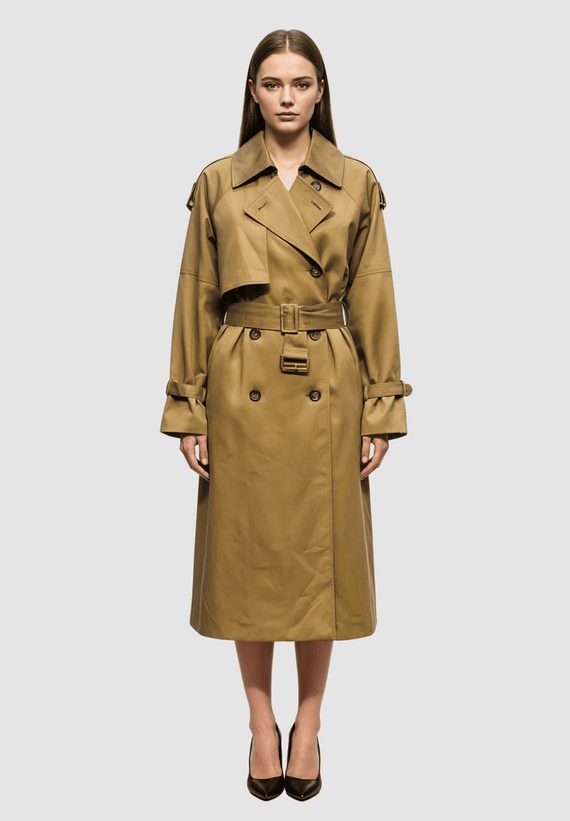 Mujer de pie, de frente, vistiendo un largo abrigo trench color marrón claro con botones dobles y cintura ceñida con cinturón, tacones negros, fondo neutro.