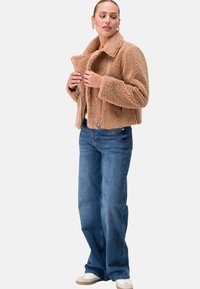 Beige, strukturierte Kunstfelljacke mit einer taillierten Passform und breitem Kragen. Kombiniert mit blauen gerade geschnittenen Jeans und weißen Sneakern.
