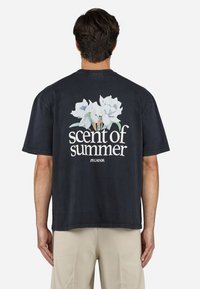 Zwart, kortarig T-shirt met een bloemenafbeelding van witte magnolia's en de tekst "geur van de zomer" in het wit. Relaxte pasvorm.
