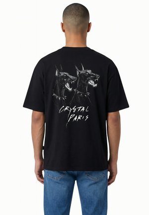 Homme portant un t-shirt noir avec un graphisme de deux chiens Doberman grognants et le texte « Crystal Paris » dans le dos, associé à un jean bleu.