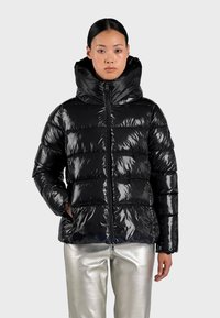 Giacca a vento nera con texture lucida e trapuntata, colletto alto e zip frontale. Abbinata a pantaloni metallici argentati.