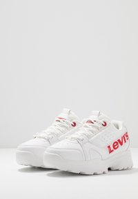 White sneakers with a textured leather upper, red logo accents, and a chunky outsole featuring prominent tread pattern.