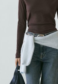 Persoon die een donker shirt met lange mouwen draagt, blauwe spijkerbroek met riem, een lichtblauwe trui om de taille gestrikt en een zwarte tas vasthoudt.