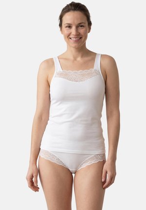 Frau lächelt und trägt ein weißes, mit Spitze besetztes Camisole und passende, mit Spitze verzierte Unterwäsche, steht vor einem schlichten, hellen Hintergrund.