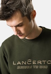Oliwkowy sweter wykonany z mieszanki bawełny; zdobią go hafty "LANCERTO" oraz "DRIVEN TO WIN" w odcieniu beżowym. Klasyczny okrągły dekolt i miękka tekstura.