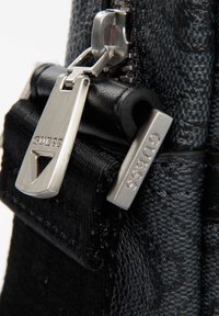 Schwarze Handtasche mit Reißverschluss, die eine strukturierte Oberfläche, silberne Beschläge und ein auffälliges "GUESS"-Logo an den Reißverschlussschiebern aufweist.