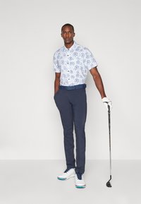 Koszula z krótkim rękawem w niebiesko-białe wzory, granatowe spodnie, białe buty golfowe z niebieskimi akcentami; trzyma kij golfowy.