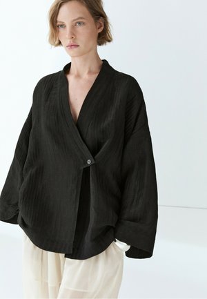 Femme aux cheveux blonds courts portant un haut noir ample style cache-cœur avec un seul bouton et un pantalon large de couleur claire devant un fond uni.