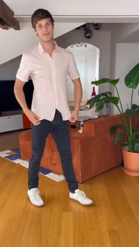 Svetlo roza kratki rokavi s gumbi, temni slim fit hlače in bele športne copate. Ozadje vključuje rastlino in oranžno kavčev. Lesena tla.