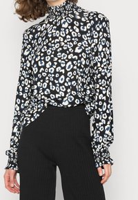 Donna che indossa una blusa a maniche lunghe e collo alto con una stampa leopardo nero, bianco e blu abbinata a pantaloni neri a costine.