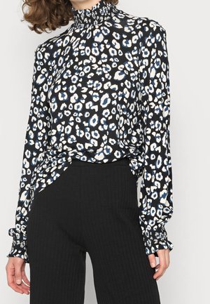 Donna che indossa una blusa a maniche lunghe e collo alto con una stampa leopardo nero, bianco e blu abbinata a pantaloni neri a costine.