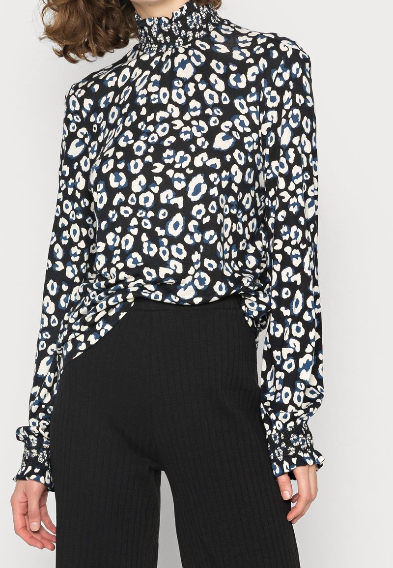 Donna che indossa una blusa a maniche lunghe e collo alto con una stampa leopardo nero, bianco e blu abbinata a pantaloni neri a costine.