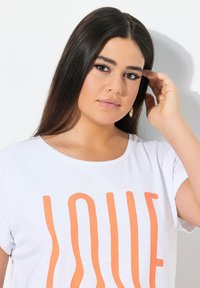 Witte t-shirt met korte mouwen en grote oranje "LOVE" opdruk. Soepele stof, casual pasvorm, ronde hals en opgerolde mouwen.