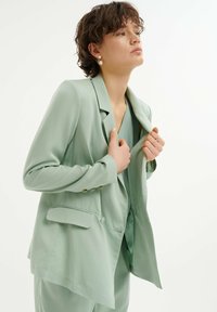 Světle zelený tailored blazer s klopovými límci, předními kapsami, zlatými knoflíky a jemnou, splývavou látkou. Nosený přes ladící top.