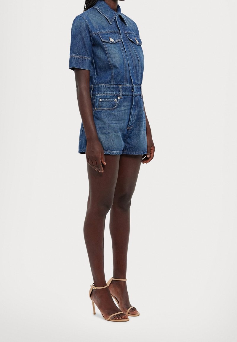 Femme portant une combinaison en denim à manches courtes avec col et poches, associée à des sandales à talons hauts beiges, debout de profil sur fond blanc.