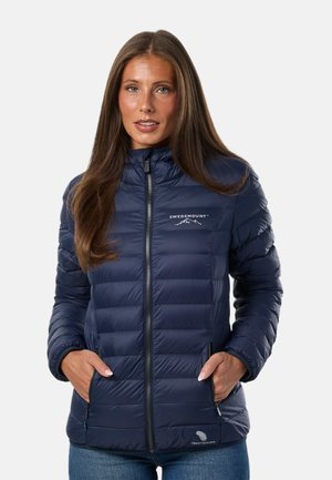 Femme portant une veste matelassée bleu marine avec poches zippées et logo Swedemount, debout les mains dans les poches devant un fond uni.