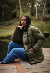 Veste matelassée vert olive avec capuche, présentant un motif matelassé et un cordon noir. Associée à un jean large bleu.