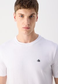 Hvid bomulds t-shirt med rund hals, korte ærmer, og et lille sort "&" logo broderet på venstre brystområde.
