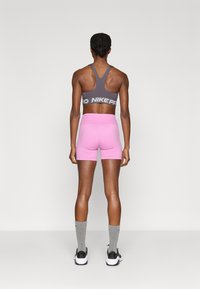 Sportski grudnjak od sive tkanine s bijelim logotipom u kombinaciji s ružičastim kratkim hlačama. Model nosi sive čarape i crne sportske tenisice.