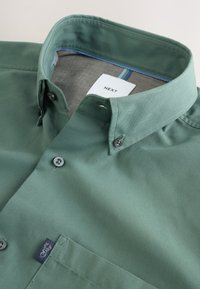 Chemise verte à boutons avec col boutonné, poche avant et étiquette de marque « NEXT » visible à l’intérieur du col.