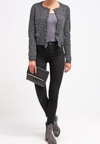 Svart, texturerad kofta med öppen front och fransade kanter, kombinerad med en grå topp, svarta skinny jeans och grå ankelstövlar; håller en svart clutch.