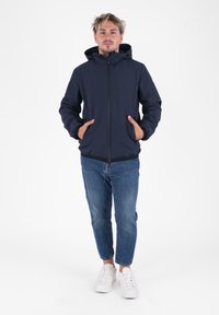 Marineblauwe hooded jack met een rits aan de voorkant, elastische manchetten en zijzakken, gecombineerd met lichtblauwe jeans en witte sneakers.