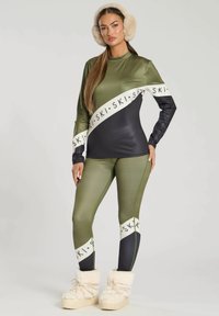 Skioutfit bestående af en olivengrøn langærmet top med sorte detaljer og logo-print, der er kombineret med matchende leggings og fyldige hvide støvler.