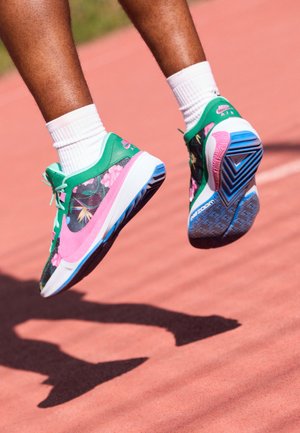 Osoba skacze na czerwonym torze, nosząc białe skarpetki oraz zielone, różowe i niebieskie, kwiatowe buty Nike Air Zoom.